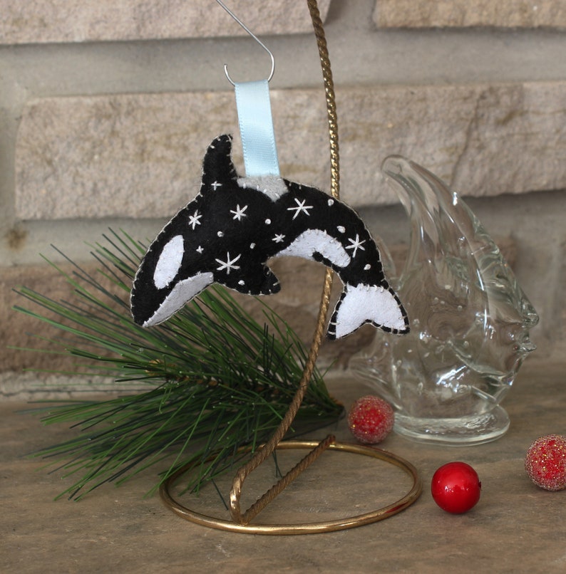 May include: Black and white felt ornament of an orca whale with white stars on a black background. The ornament is hanging from a blue ribbon on a gold metal stand.