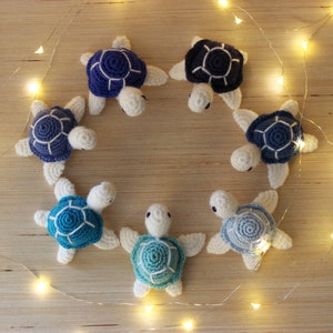 May include: Seven crocheted turtle toys arranged in a circle on a wooden surface. The turtles are white with blue and turquoise shells. The turtles are arranged in a circle with their heads facing inwards.