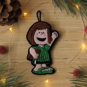 May include: A felt Christmas ornament depicting a cartoon character in a green dress and sandals, with brown hair and a waving hand. The ornament has a black loop for hanging. The background includes pine needles, berries, and fairy lights.