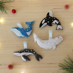 May include: Four felt whale ornaments with a snowflake pattern. The whales are blue, white, black, and gray. They are hanging from white ribbon.