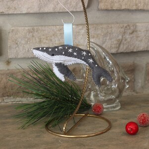 May include: A gray felt whale ornament with white embroidered stars, hanging from a blue ribbon on a gold stand. The whale is a Christmas ornament.