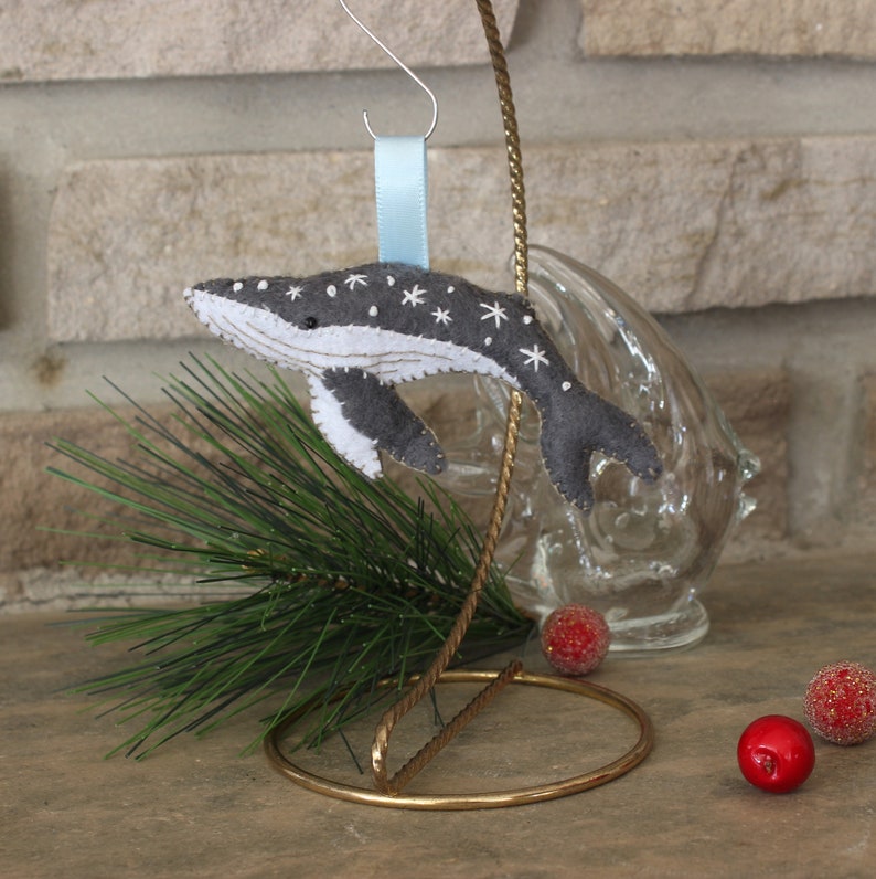 May include: A gray felt whale ornament with white embroidered stars, hanging from a blue ribbon on a gold stand. The whale is a Christmas ornament.