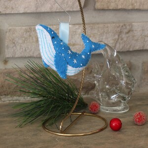 May include: Blue felt whale ornament with white stitching and snowflake details. The ornament is hanging from a gold wire stand with a blue ribbon.