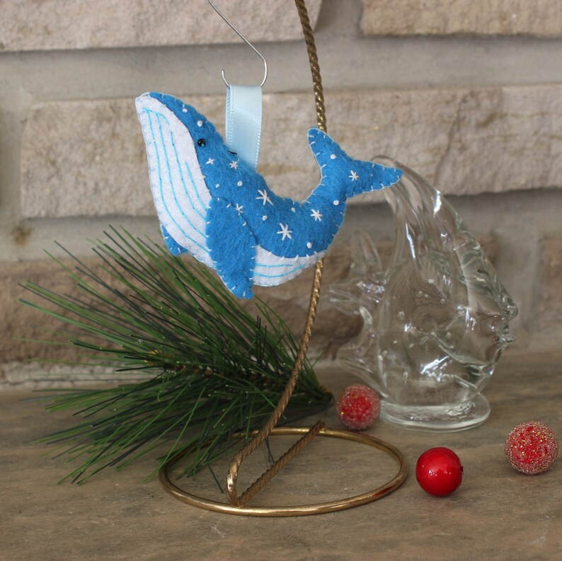 May include: Blue felt whale ornament with white stitching and snowflake details. The ornament is hanging from a gold wire stand with a blue ribbon.