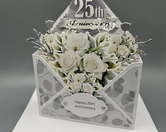 May include: A 3D greeting card shaped like an envelope featuring a bouquet of white roses and green leaves. The card displays the text 25th Anniversary at the top and Happy 25th Anniversary on a white oval label at the bottom with small silver accents.