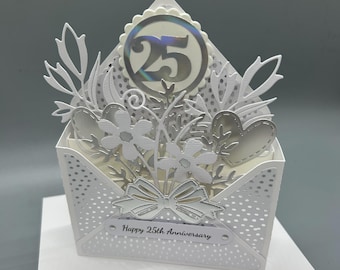 May include: A white papercraft anniversary card with a die-cut envelope design. The card features a silver foil number 25, floral embellishments, and the text "Happy 25th Anniversary". The card is decorated with delicate cut-out details.