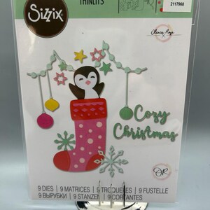 May include: Sizzix Thinlits die set featuring a penguin in a Christmas stocking, with stars, snowflakes, and the words "Cozy Christmas." The set includes 9 dies and is designed for paper crafts.