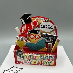 May include: A red and white graduation-themed pop-up card with a cartoon student reading a book. The card includes a graduation cap, the year 2026, and the words "Graduation Day". Other elements include a pencil, trophy, and medical symbol.