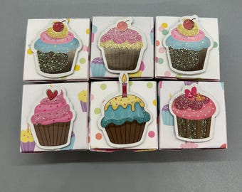 May include: Six small white boxes with cupcake stickers. Each cupcake has a different colored frosting, including pink, blue, and yellow, with glitter accents and a cherry on top. One cupcake has a lit candle.
