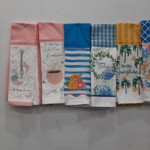 May include: A collection of decorative kitchen towels with various designs. One towel features a guitar and the text "I will always love you." Other towels display coffee cups, floral patterns, and the phrases "Happy Spring" and "hello sunshine."