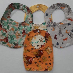 Op de afbeelding: Drie kleurrijke baby-slabbetjes met een Lion King-thema. De slabbetjes hebben verschillende patronen met leeuwen, junglebegroeiing en de woorden "Hakuna Matata".