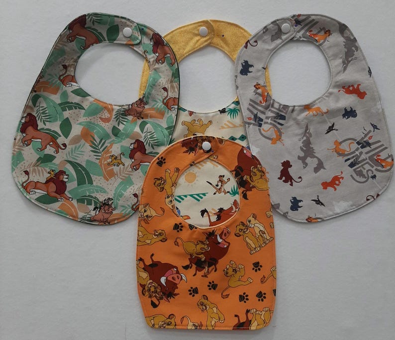 Op de afbeelding: Drie kleurrijke baby-slabbetjes met een Lion King-thema. De slabbetjes hebben verschillende patronen met leeuwen, junglebegroeiing en de woorden "Hakuna Matata".