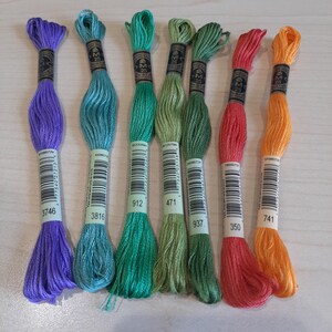 May include: A collection of embroidery floss in various colors, including purple, teal, green, coral, and orange. Each skein is labeled with a number and brand information. The floss is neatly arranged, showcasing the vibrant hues.