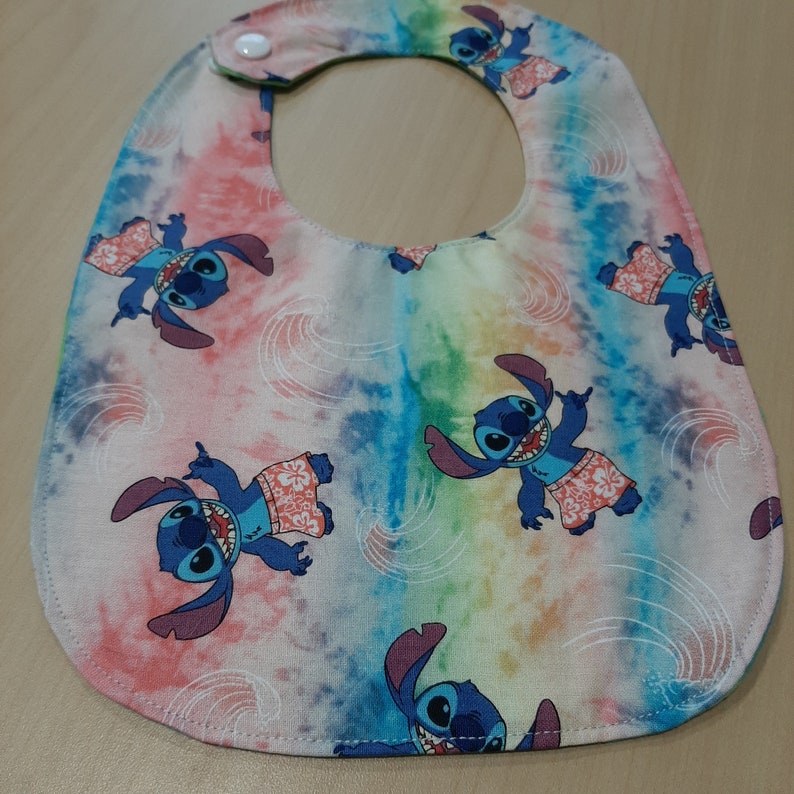 Puede incluir: Babero para beb&eacute; con un colorido estampado tie-dye con personajes de dibujos animados azules con camisas hawaianas. El babero tiene un cierre de bot&oacute;n a presi&oacute;n en el cuello.