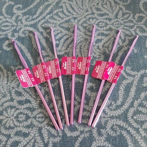 May include: A collection of six pink aluminum crochet hooks, each with a label that reads "Susan Bates Aluminum/Aluminium 4.0 G-6". The hooks are arranged in a fan shape on a blue patterned surface.