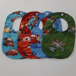 May include: Four baby bibs with snap closures. The bibs feature colorful cartoon car and airplane designs on a light blue, red, blue, and green background. The bibs are made of soft fabric and are designed to catch spills.