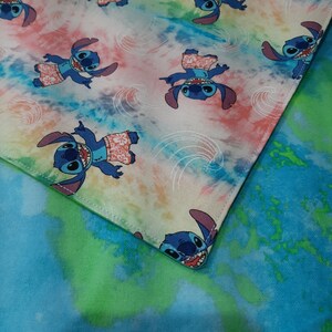 Puede incluir: Una tela cuadrada con estampado tie-dye que presenta al personaje Stitch de Lilo & Stitch. Stitch se muestra en varias poses, usando pantalones cortos hawaianos. El fondo tiene un efecto de acuarela con tonos azules, rosas y amarillos.