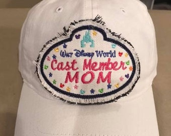May include: White baseball cap with a patch that reads "Our Disney World Cast Member MOM." The patch has a blue border with colorful hearts and stars. The text "MOM" is in pink.