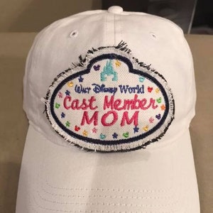 May include: White baseball cap with a patch that reads "Our Disney World Cast Member MOM." The patch has a blue border with colorful hearts and stars. The text "MOM" is in pink.