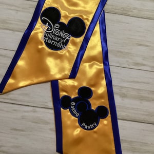 May include: A gold satin graduation sash with blue trim and embroidered Mickey Mouse silhouettes. The sash features the text "Disney Culinary Internship" and "Baking Pastry".