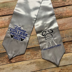 K&ouml;nnte beinhalten: Silberne Abschluss-Sch&auml;rpe mit blauer und schwarzer Stickerei "Disney College Program 2023" und Mickey Mouse Ohren. Die andere Seite zeigt das Hollywood Studios Logo.