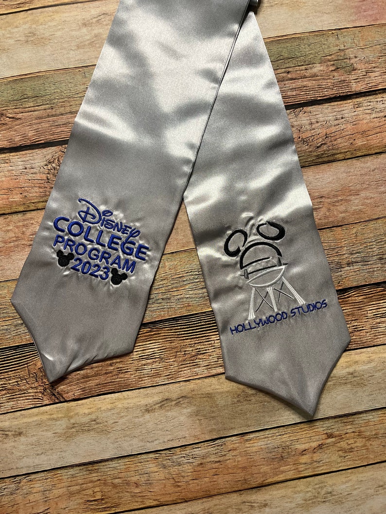 K&ouml;nnte beinhalten: Silberne Abschluss-Sch&auml;rpe mit blauer und schwarzer Stickerei "Disney College Program 2023" und Mickey Mouse Ohren. Die andere Seite zeigt das Hollywood Studios Logo.
