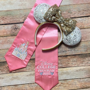 K&ouml;nnte beinhalten: Rosa Abschluss-Satin-Sch&auml;rpe mit gesticktem Schloss und Text "Disney College Program 2023". Eine goldene Pailletten-Schleife und ein Ohren-Stirnband mit wei&szlig;en und goldenen Akzenten.