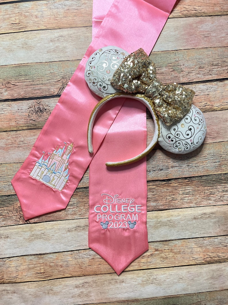 K&ouml;nnte beinhalten: Rosa Abschluss-Satin-Sch&auml;rpe mit gesticktem Schloss und Text "Disney College Program 2023". Eine goldene Pailletten-Schleife und ein Ohren-Stirnband mit wei&szlig;en und goldenen Akzenten.