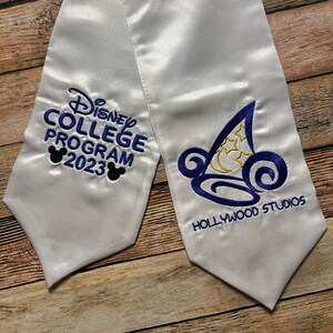K&ouml;nnte beinhalten: Wei&szlig;e Abschluss-Sch&auml;rpe aus Satin mit blauer Stickerei. Die Sch&auml;rpe tr&auml;gt den Text "Disney College Program 2023" und "Hollywood Studios", sowie Mickey Mouse Silhouetten und ein stilisiertes Hut-Design.