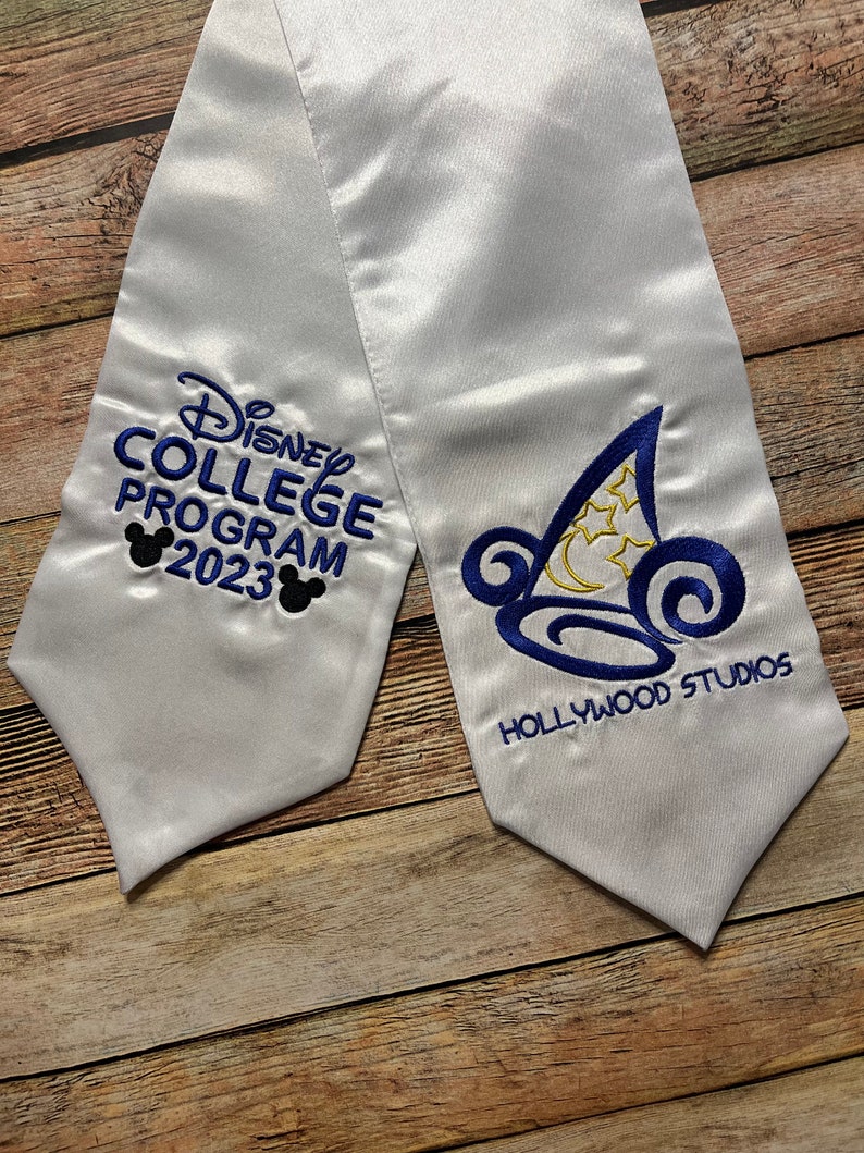 K&ouml;nnte beinhalten: Wei&szlig;e Abschluss-Sch&auml;rpe aus Satin mit blauer Stickerei. Die Sch&auml;rpe tr&auml;gt den Text "Disney College Program 2023" und "Hollywood Studios", sowie Mickey Mouse Silhouetten und ein stilisiertes Hut-Design.