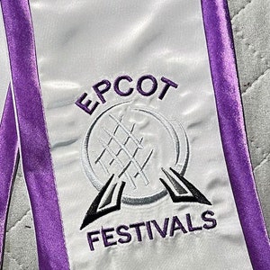 K&ouml;nnte beinhalten: Abschluss-Sch&auml;rpe aus wei&szlig;em Satin mit lila Besatz. Die Sch&auml;rpe zeigt den gestickten Text "EPCOT FESTIVALS" in Lila und Schwarz sowie ein stilisiertes Logo. Die Sch&auml;rpe ist f&uuml;r eine Abschlussfeier bestimmt.