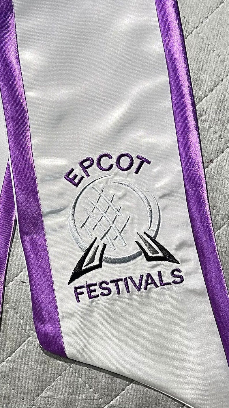K&ouml;nnte beinhalten: Abschluss-Sch&auml;rpe aus wei&szlig;em Satin mit lila Besatz. Die Sch&auml;rpe zeigt den gestickten Text "EPCOT FESTIVALS" in Lila und Schwarz sowie ein stilisiertes Logo. Die Sch&auml;rpe ist f&uuml;r eine Abschlussfeier bestimmt.
