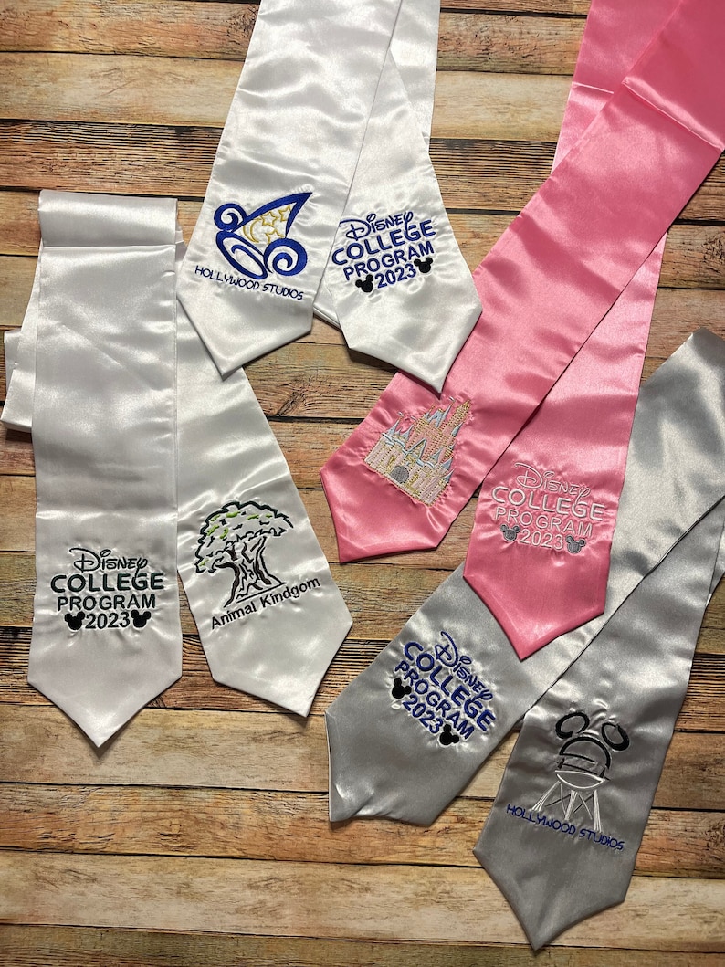 K&ouml;nnte beinhalten: Abschlussstolen aus Satin in Wei&szlig;, Rosa und Silber, bestickt mit Disney College Program 2023 Logos und Park-Icons. Die Stolen zeigen Designs f&uuml;r Hollywood Studios und Animal Kingdom.