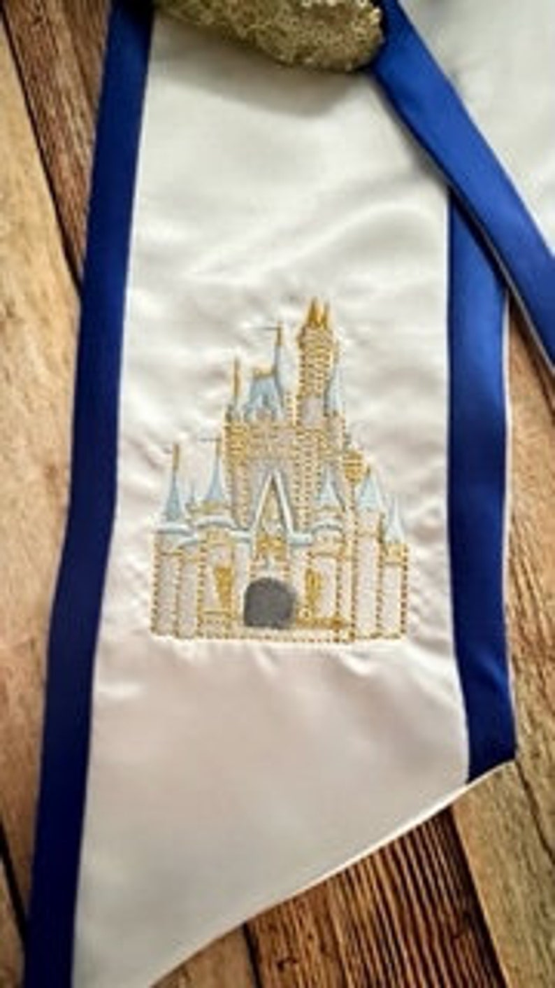 K&ouml;nnte beinhalten: Wei&szlig;es Satin-Sch&auml;rpe mit blauem Rand und goldener Pailletten-Oberseite. Die Sch&auml;rpe zeigt ein gesticktes Bild eines Schlosses mit goldenen, blauen und grauen Details. Die Sch&auml;rpe wird auf einer Holzoberfl&auml;che pr&auml;sentiert.