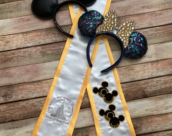 Könnte beinhalten: Weiße und goldene Abschluss-Schärpe mit dem Text "The World's Most Magical Celebration" und "Walt Disney World". Außerdem schwarze Mickey Mouse Ohren-Stirnbänder mit Pailletten und goldenen Akzenten.