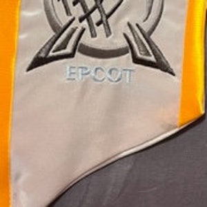K&ouml;nnte beinhalten: Eine wei&szlig;e Satin-Abschluss-Sch&auml;rpe mit grau gesticktem EPCOT-Logo. Die Sch&auml;rpe hat einen leuchtend gelben Rand. Das EPCOT-Logo zeigt ein stilisiertes Globus-Design mit dem Wort "EPCOT" darunter.