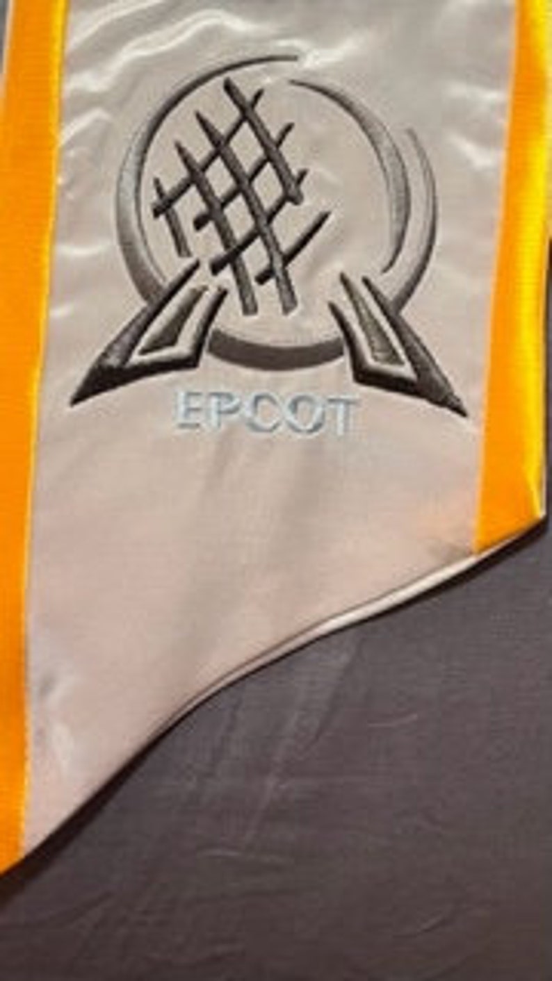 K&ouml;nnte beinhalten: Eine wei&szlig;e Satin-Abschluss-Sch&auml;rpe mit grau gesticktem EPCOT-Logo. Die Sch&auml;rpe hat einen leuchtend gelben Rand. Das EPCOT-Logo zeigt ein stilisiertes Globus-Design mit dem Wort "EPCOT" darunter.
