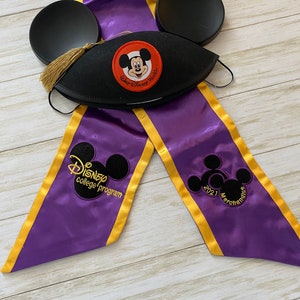 May include: A black graduation cap with a Mickey Mouse patch and gold tassel, paired with a purple and gold sash. The sash features the text "Disney college program" and "2021 Merchandise". Black mouse ear headwear is also included.