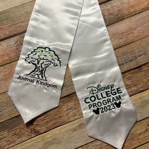 K&ouml;nnte beinhalten: Wei&szlig;e Abschluss-Satin-Sch&auml;rpe mit gesticktem Text und Grafiken. Auf der einen Seite steht "Animal Kindgom" mit einer Baumgrafik. Auf der anderen Seite steht "Disney College Program 2023" mit Mickey-Mouse-Silhouetten.