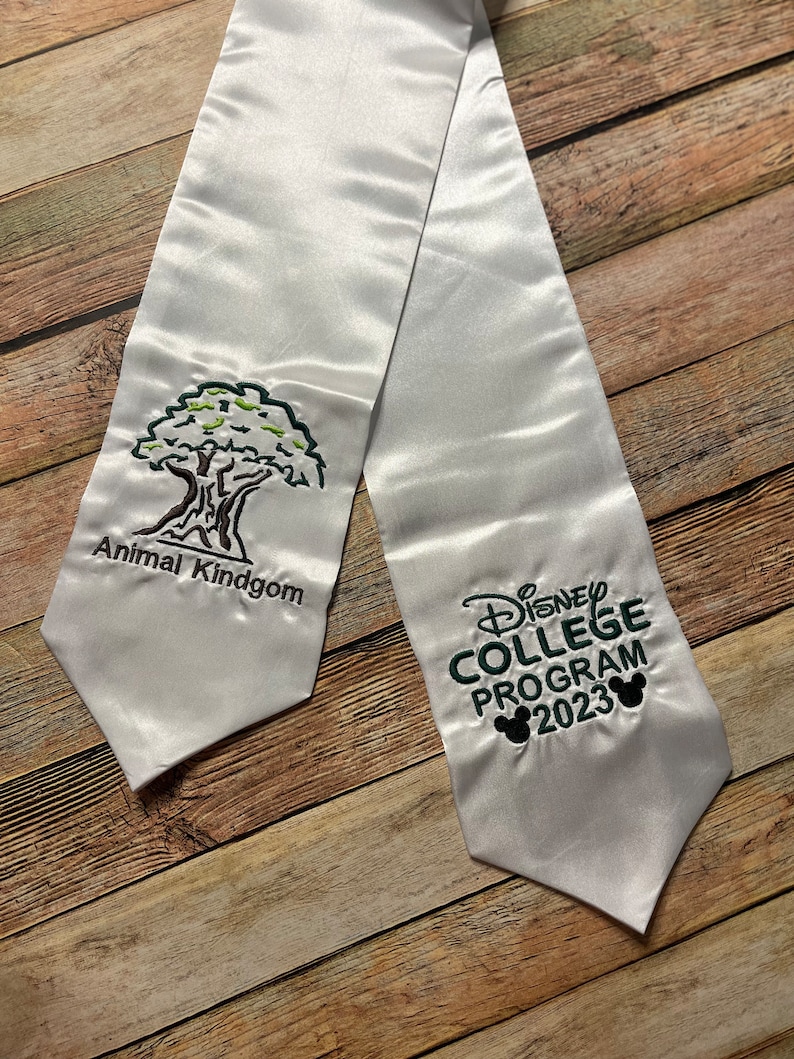 K&ouml;nnte beinhalten: Wei&szlig;e Abschluss-Satin-Sch&auml;rpe mit gesticktem Text und Grafiken. Auf der einen Seite steht "Animal Kindgom" mit einer Baumgrafik. Auf der anderen Seite steht "Disney College Program 2023" mit Mickey-Mouse-Silhouetten.
