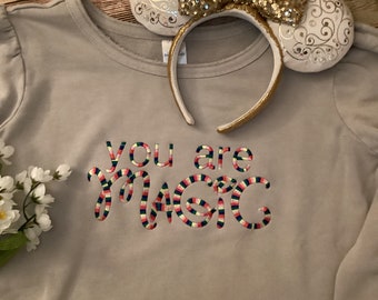 Könnte beinhalten: Ein hellgraues Sweatshirt mit den gestickten Worten "you are MAGIC" in bunten Streifen. Ein goldfarbenes Pailletten-Schleifen-Stirnband mit weißen und goldenen Ohren liegt über dem Shirt. Weiße Blumen befinden sich in der linken unteren Ecke.