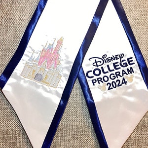 K&ouml;nnte beinhalten: Wei&szlig;e Abschluss-Sch&auml;rpe mit marineblauer Einfassung. Mit einem gestickten rosa-goldenen Schloss und dem Text "Disney College Program 2024" in Marineblau mit Mickey Mouse-K&ouml;pfen. Ein festliches Accessoire.