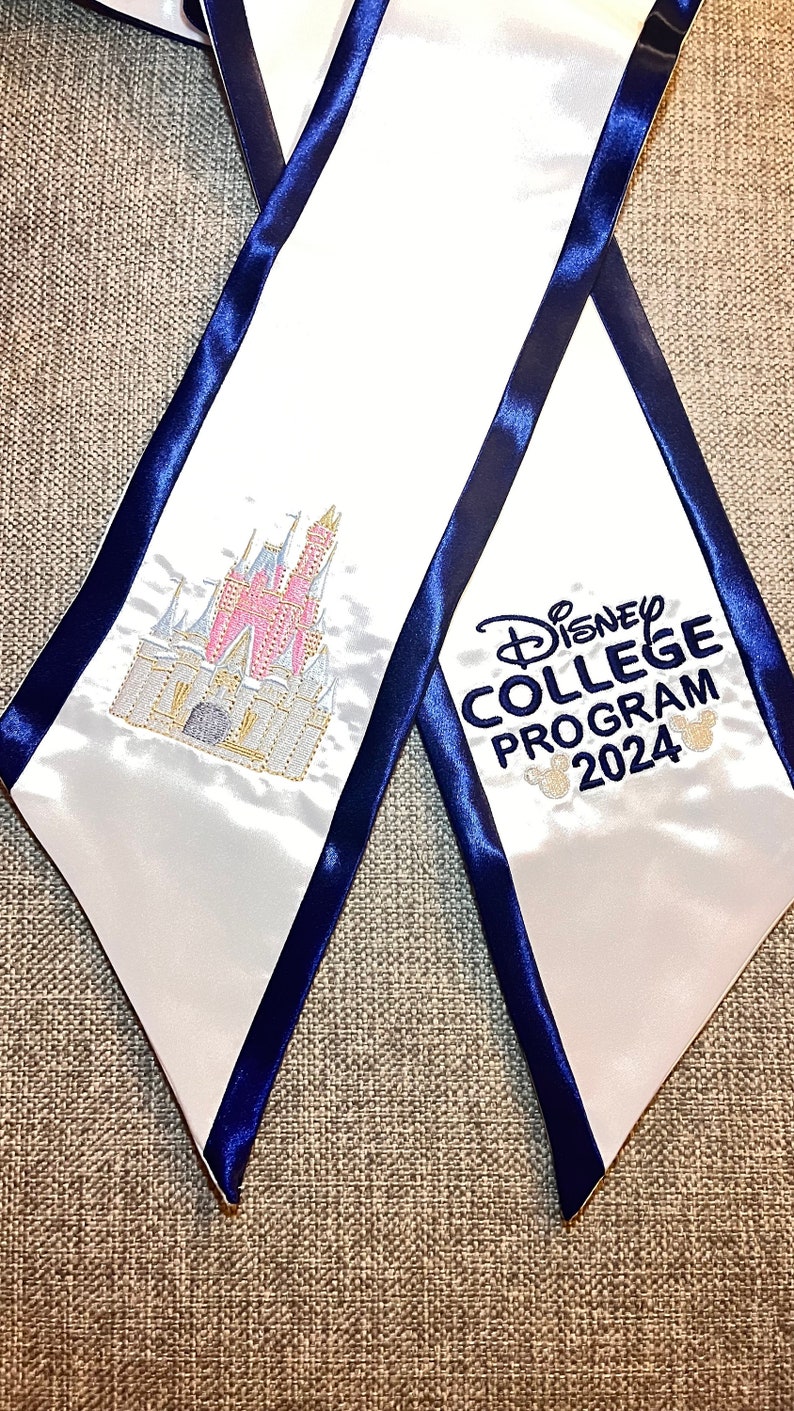 K&ouml;nnte beinhalten: Wei&szlig;e Abschluss-Sch&auml;rpe mit marineblauer Einfassung. Mit einem gestickten rosa-goldenen Schloss und dem Text "Disney College Program 2024" in Marineblau mit Mickey Mouse-K&ouml;pfen. Ein festliches Accessoire.