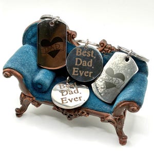 Peut inclure: Plusieurs porte-clés en argent gravés des inscriptions « Best Dad Ever » et « Dad ». Ils sont présentés sur un petit canapé en velours bleu avec un cadre en bois marron, un objet décoratif.