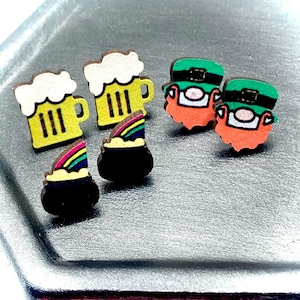 Puede incluir: Pendientes de madera con temática del Día de San Patricio. Los pendientes incluyen diseños de jarras de cerveza, duendes y ollas de oro con arcoíris. Los pendientes están sobre una bandeja plateada.