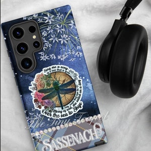 May include: A blue phone case with a dragonfly design and the text "Sing me a song of a lass that is gone". The case features a floral and plaid pattern, with the word "Sassenach" displayed prominently. A pair of black headphones rests nearby.