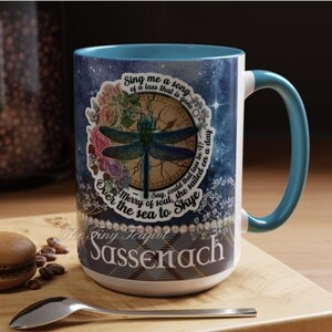 May include: A ceramic mug with a blue interior and handle, featuring a dragonfly design with floral accents and text. The mug has the words "Sassenach" and other text. The mug is on a wooden surface with a spoon and a pastry.