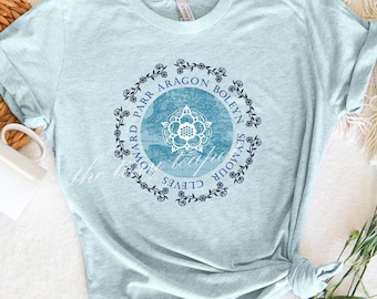 May include: Light blue t-shirt with a blue and white graphic of a Tudor rose surrounded by a wreath of flowers and the names of Henry VIII's wives: Catherine of Aragon, Anne Boleyn, Jane Seymour, Anne of Cleves, Catherine Howard, Catherine Parr.