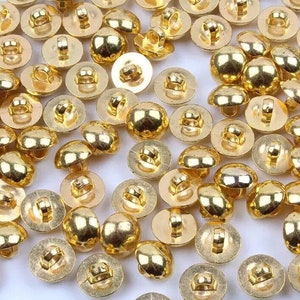 Könnte beinhalten: Eine Ansammlung glänzender, goldfarbener Knöpfe. Die Knöpfe haben eine abgerundete, gewölbte Form mit einer Schlaufe auf der Rückseite zum Annähen. Die Knöpfe sind über die Oberfläche verstreut und zeigen ihre reflektierende, metallische Oberfläche. Diese Knöpfe eignen sich für verschiedene Nähprojekte.
