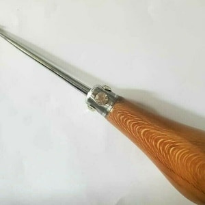 Könnte beinhalten: Ein langes, dünnes, silbernes Werkzeug mit einem Holzgriff. Der Griff ist hellbraun mit einer Holzmaserung. Das Werkzeug hat eine spitze Spitze und einen Metallring in der Nähe des Griffs.